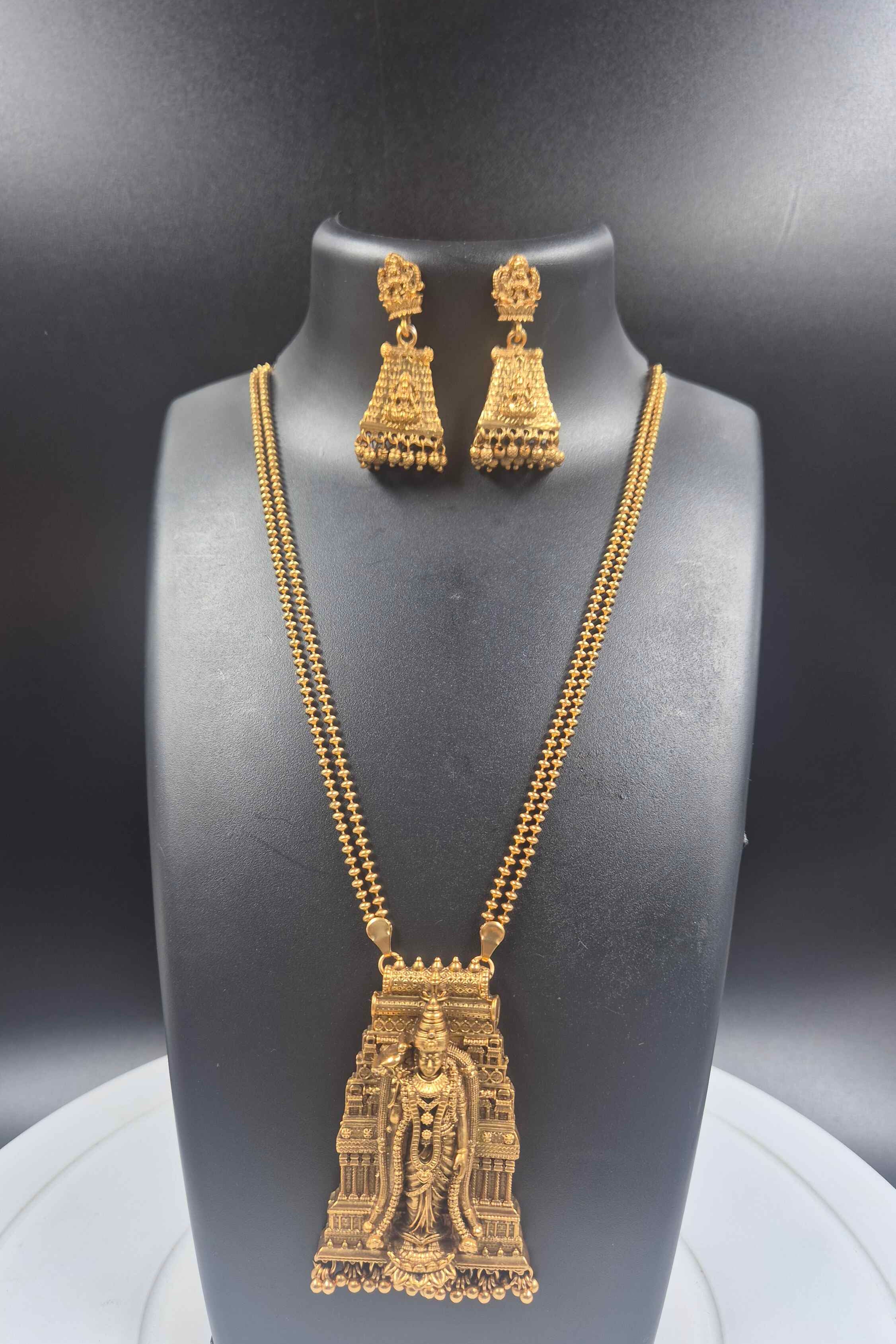 Gold-toned jewelry set displayed on a black jewelry bust, featuring a necklace with a detailed temple tower pendant and matching drop earrings, set against a dark gray background.